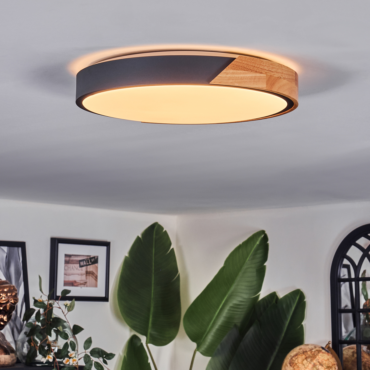 Eine moderne Deckenlampe mit Holz- und schwarzen Akzenten ist zu sehen. Darunter sind große grüne Blätter einer Pflanze zu sehen.