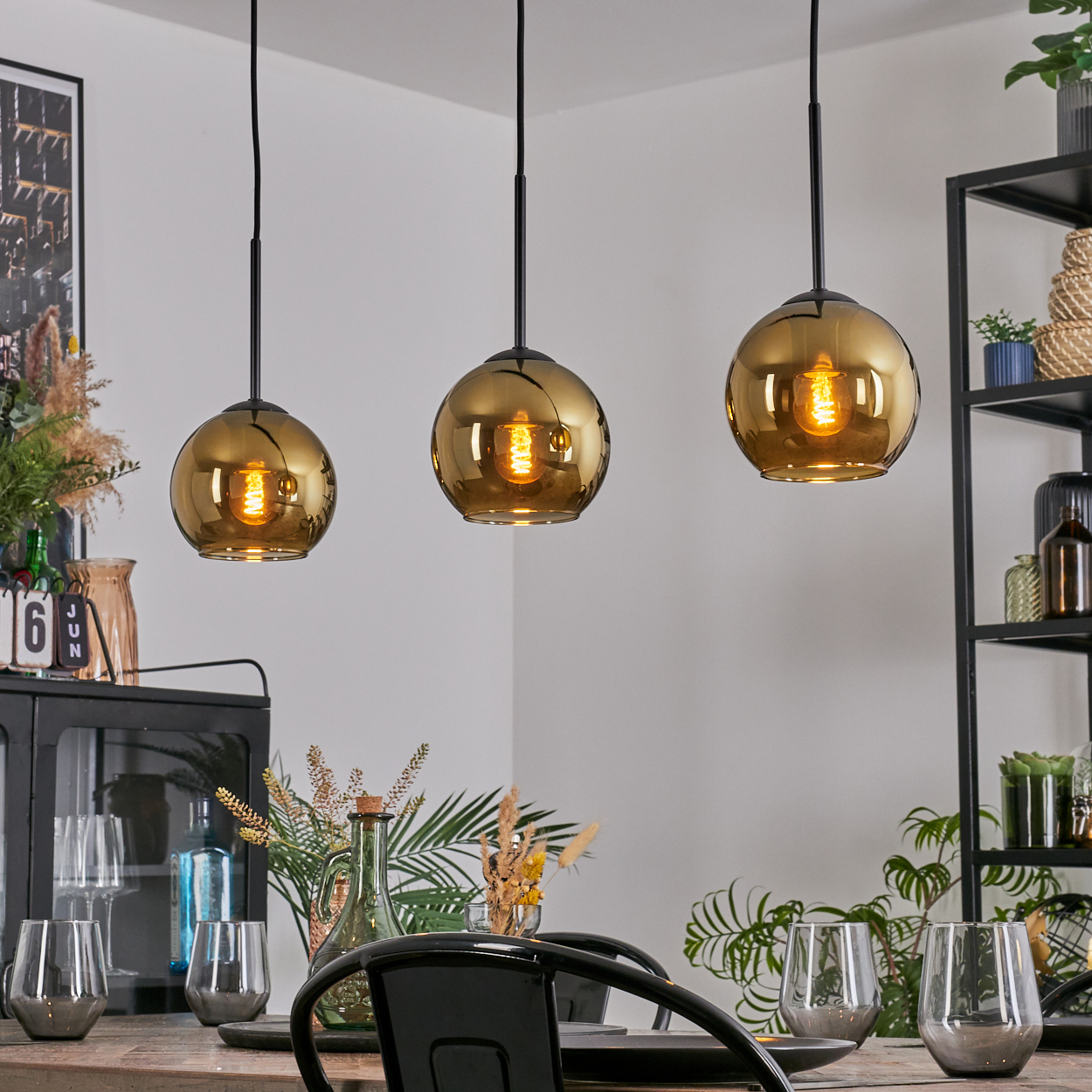Drei goldene Pendelleuchten hängen über einem Esstisch mit Gläsern und Pflanzen. Ein schwarzes Regal und ein Schrank sind im Hintergrund.