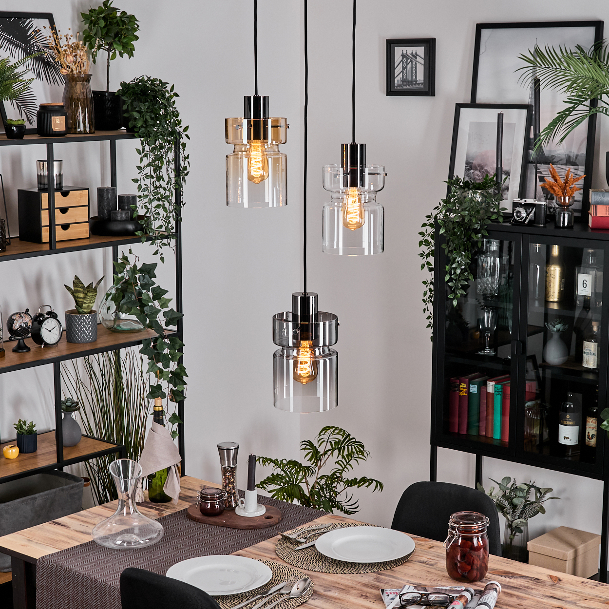 Esszimmer mit Holztisch, drei Hängelampen, Regalen mit Deko und Pflanzen, Tellern, Glas und schwarzen Stühlen.
