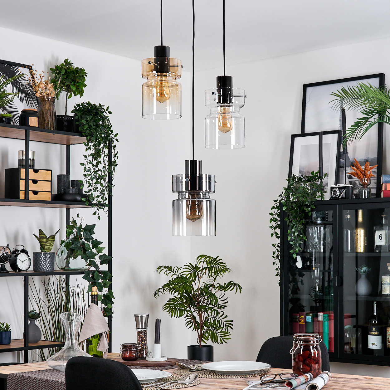 Esszimmer mit drei Hängelampen, Pflanzen, schwarzen Regalen, gedecktem Tisch und einem schwarzen Schrank. Ein Glas mit roten Früchten.