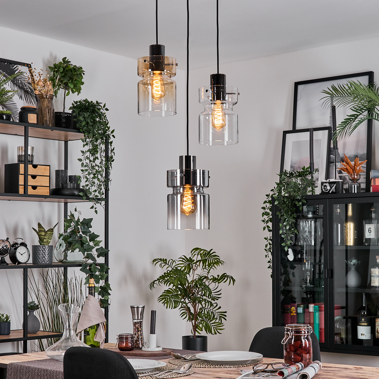 Esszimmer mit drei Hängelampen, Pflanzen, einem gedeckten Tisch und einem schwarzen Schrank mit Dekoration.