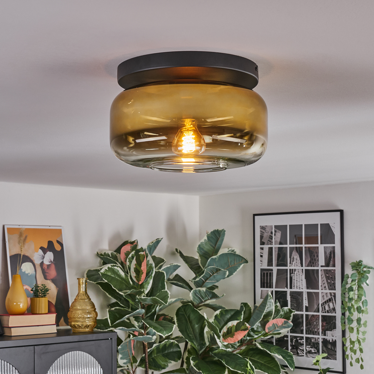 Eine moderne Deckenlampe mit Glasschirm und sichtbarer Glühbirne. Grüne Pflanzen und Kunstwerke sind im Hintergrund.