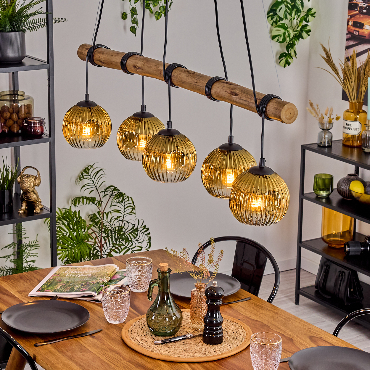 Esszimmer mit Holztisch. Sechs Hängelampen. Pflanzen und Regale im Hintergrund. Tisch gedeckt mit Tellern, Gläsern und Dekoration.