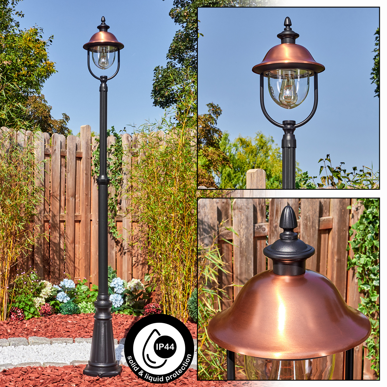 Eine schwarze Außenlampe mit kupferfarbenem Aufsatz. Sie steht in einem Garten mit Holzzaun und Pflanzen im Hintergrund.