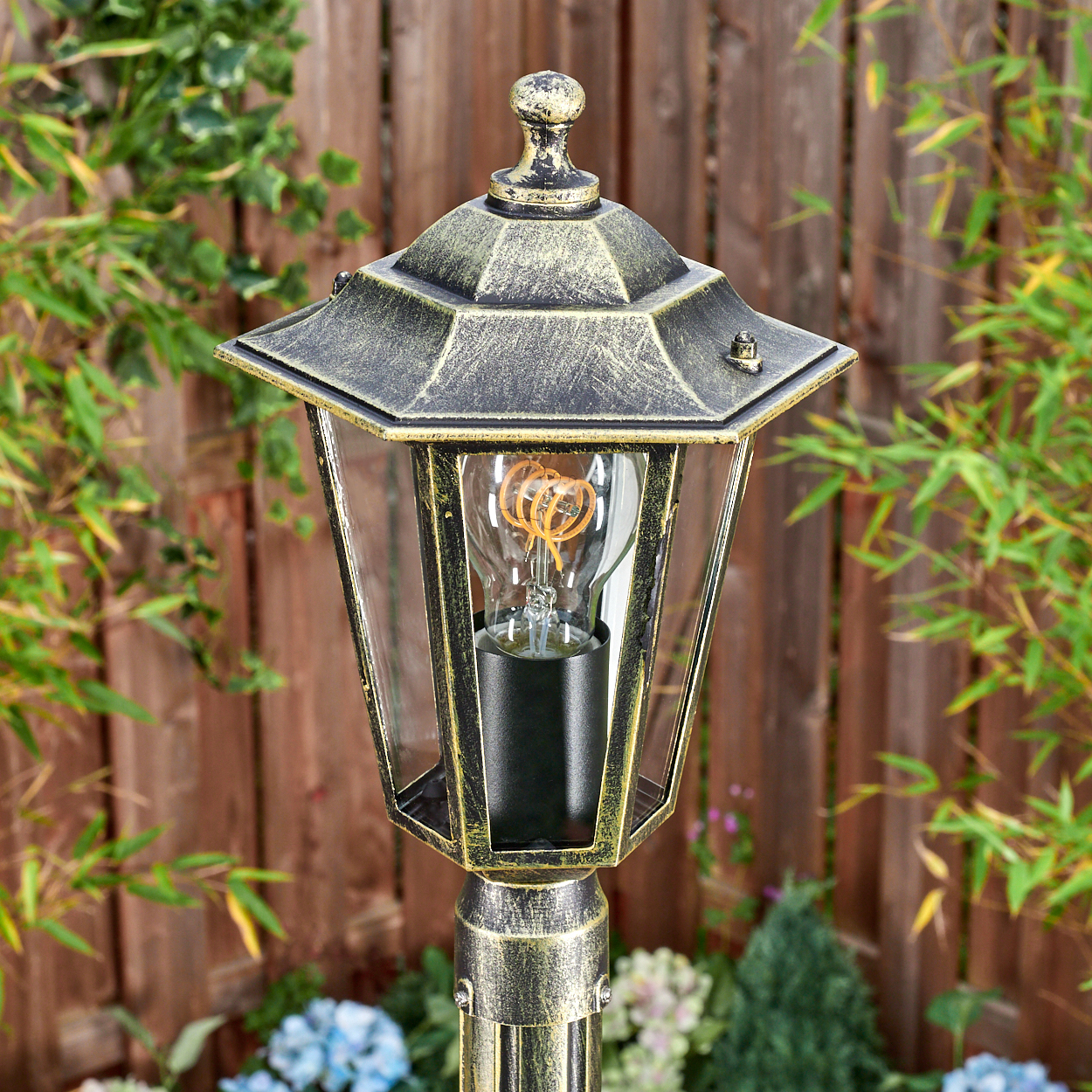 Eine verzierte Außenlaterne mit dekorativer Spitze, Glaseinfassung und Glühbirne. Sie befindet sich in einem Garten mit Pflanzen und einem Zaun.