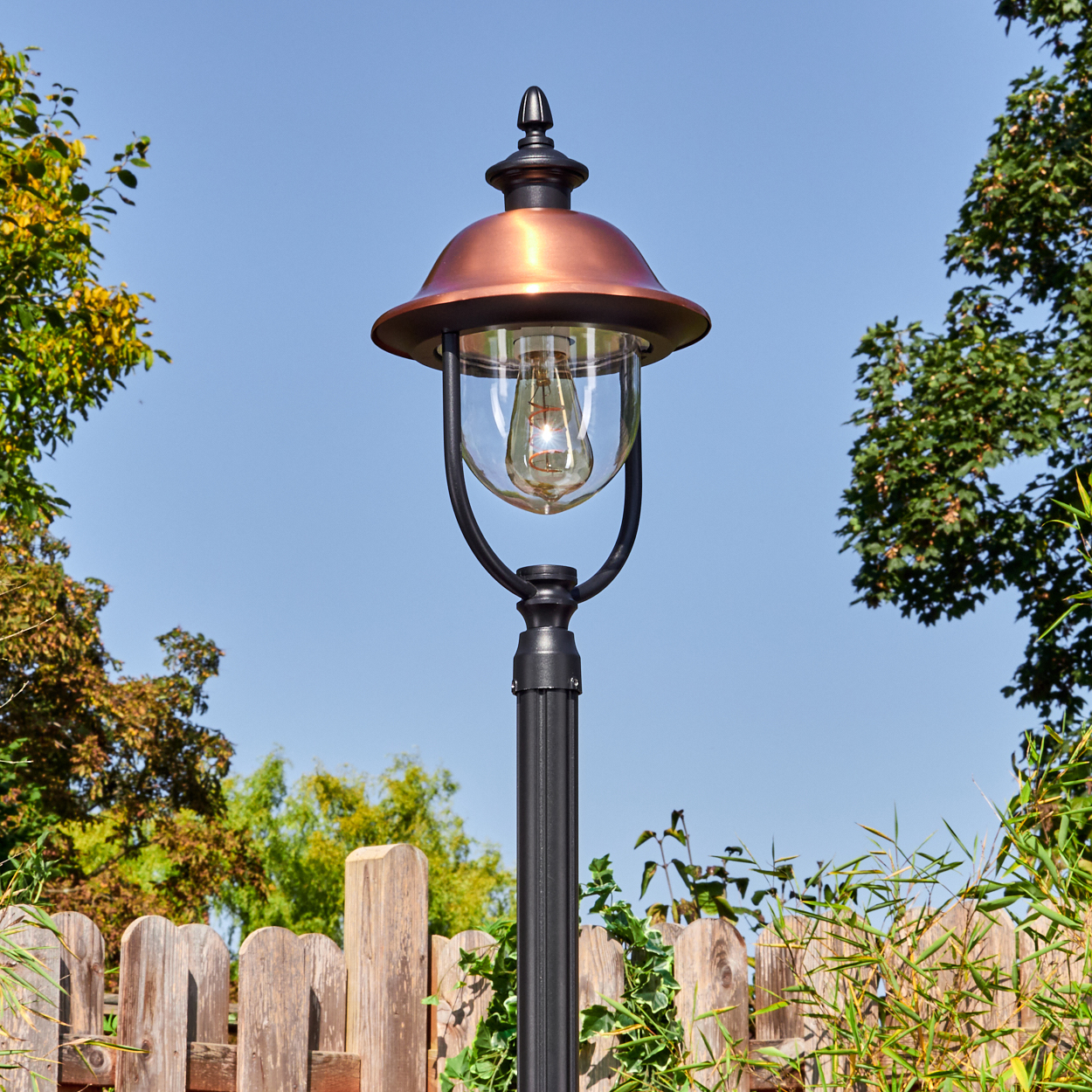 Eine dekorative Außenlampe mit kupferfarbenem Schirm und klarem Glas. Sie steht auf einer schwarzen Stange mit Holzzaun und Grün im Hintergrund.