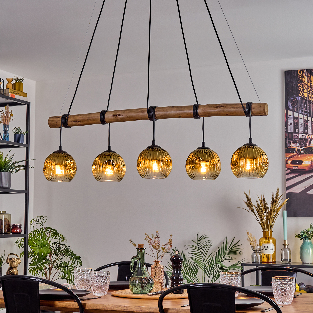Ein Esszimmer mit Holztisch, schwarzen Stühlen und Kronleuchter mit goldenen Glaslampen. Ein Gemälde und Pflanzen im Hintergrund.
