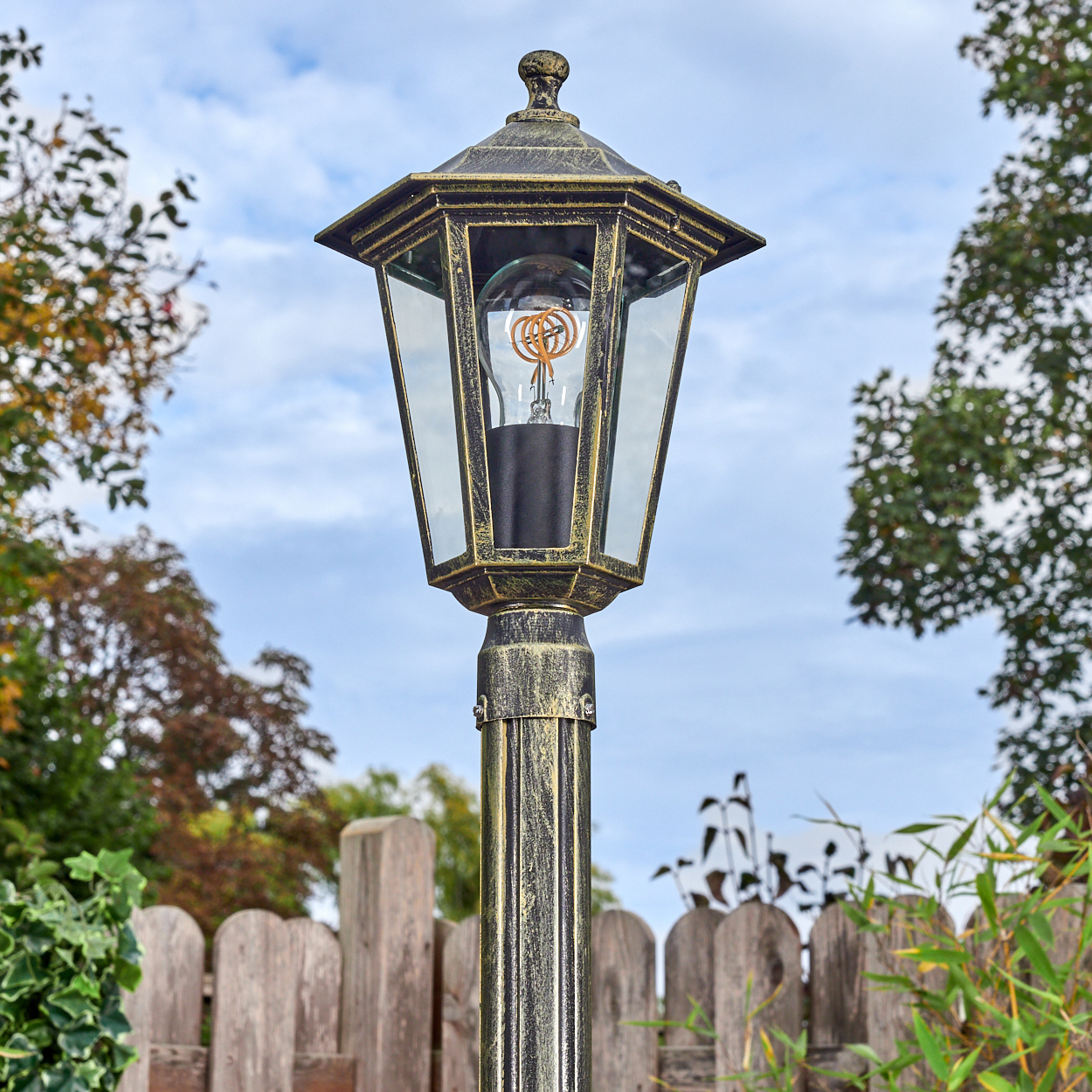 Eine verzierte, bronzefarbene Laterne mit einer glaseingeschlossenen Glühbirne steht vor einem Holzzaun und Grün, mit einem bewölkten Himmel im Hintergrund.
