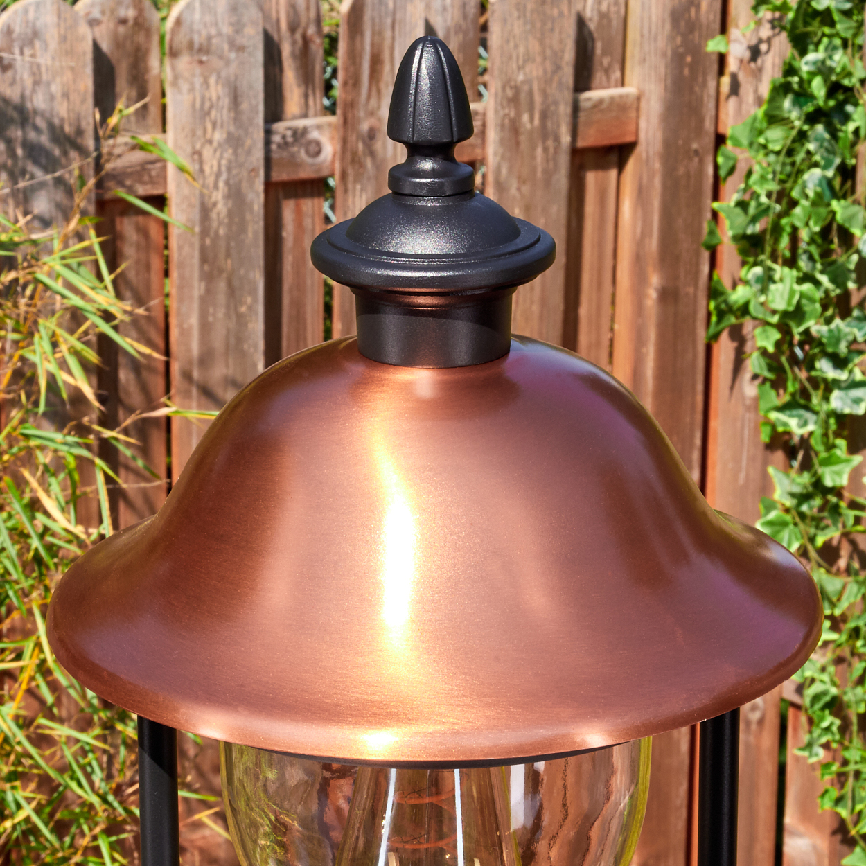 Eine kupferfarbene Außenlampe mit schwarzer Oberseite. Im Hintergrund ein Holzzaun und grüne Pflanzen.