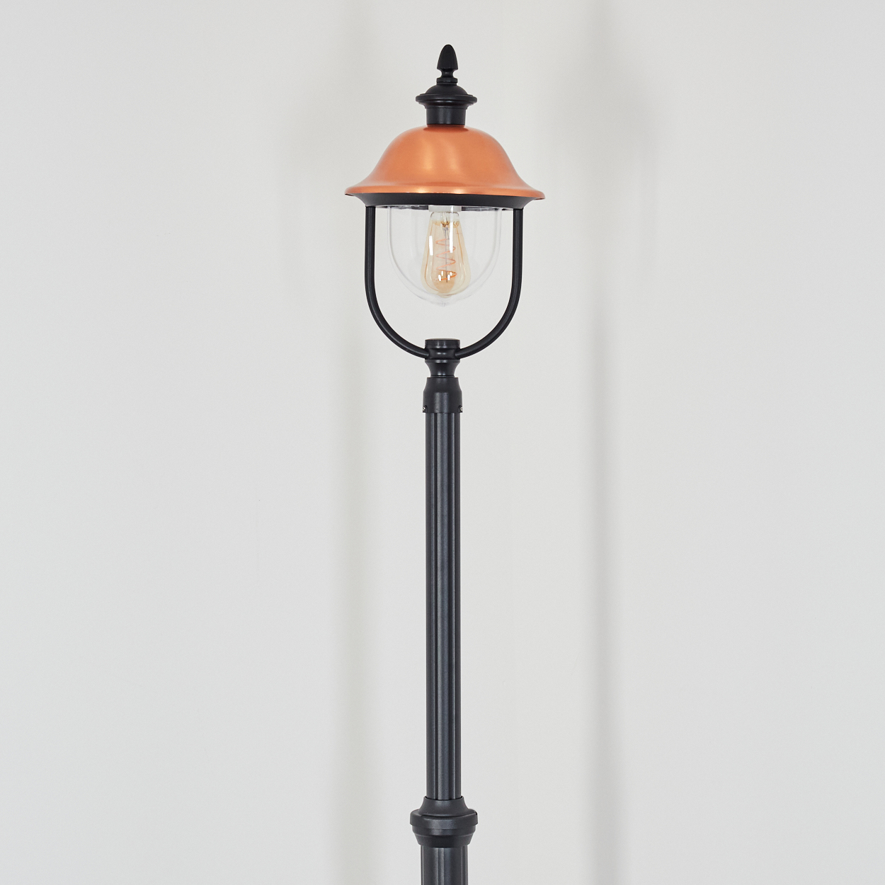 Eine schwarze und kupferfarbene Laterne vor einer weißen Wand. Die Lampe hat ein Glasgehäuse und eine dekorative Spitze.