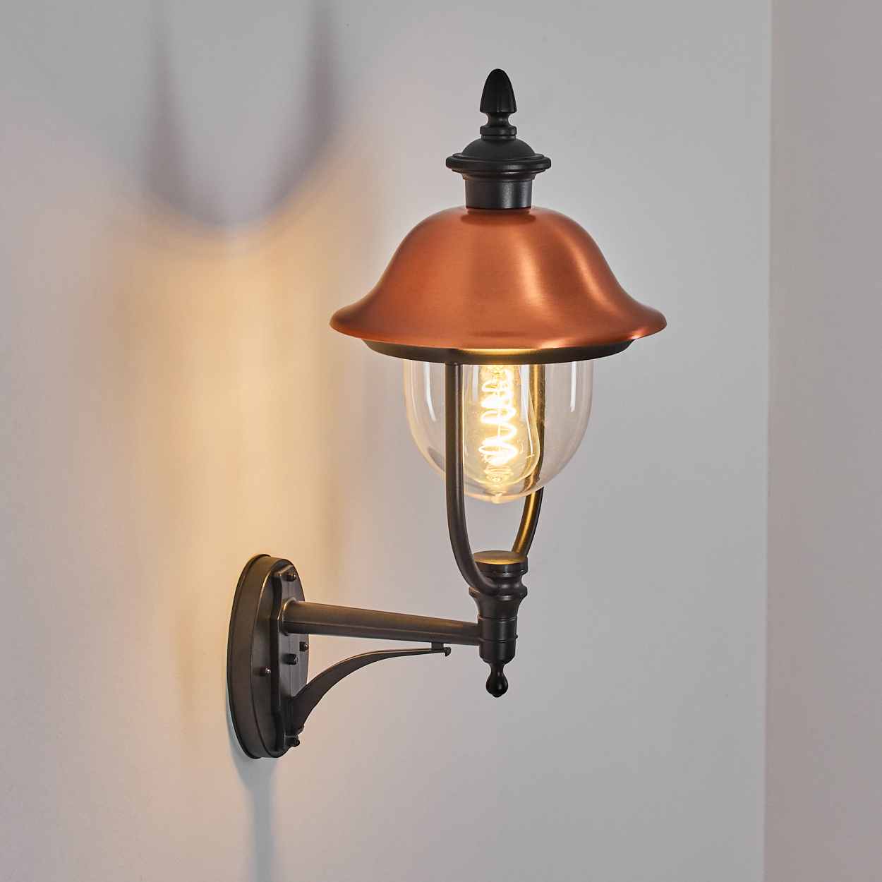 Eine wandmontierte Außenlampe aus Kupfer und Schwarz mit einem klaren Glaskolben und einer leuchtenden Glühbirne, die ein warmes Licht wirft.