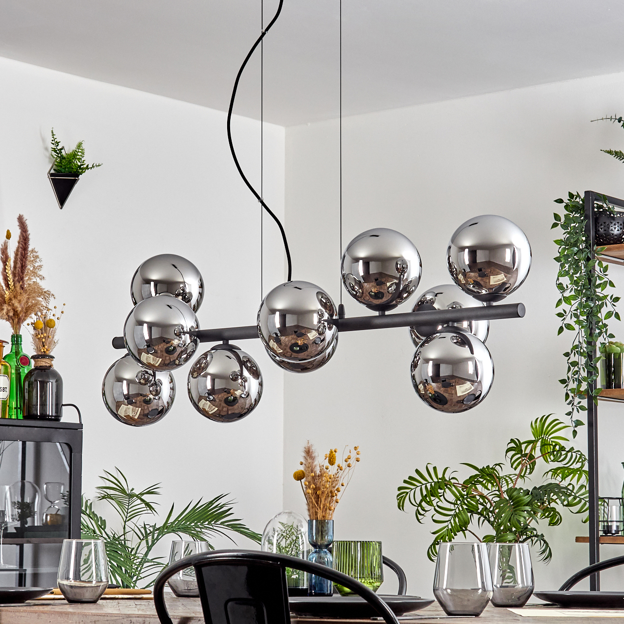 Ein modernes Esszimmer mit einem schwarzen Kronleuchter aus reflektierenden Kugeln. Tisch mit Gläsern und schwarzen Tellern gedeckt. Pflanzen und Dekoration sind ebenfalls vorhanden.