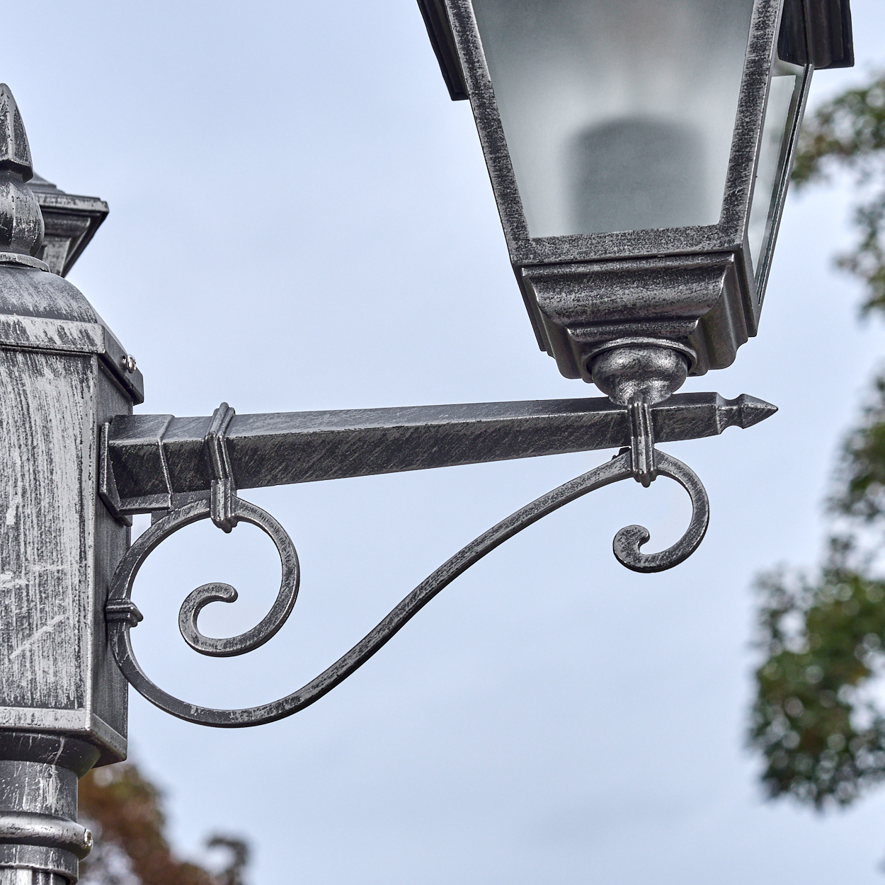 Nahaufnahme einer dekorativen, verzierten Straßenlaterne. Sie ist silberfarben mit Glasschirm und einem Schnörkeldesign. Der Himmel ist hellblau.
