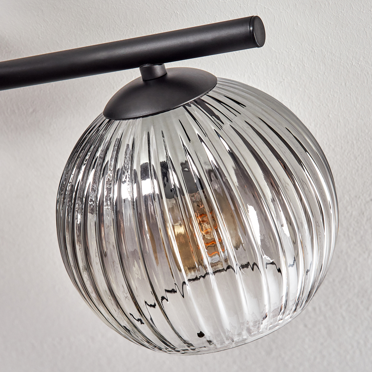Nahaufnahme einer modernen Leuchte. Sie hat einen gerippten, runden Glasschirm und einen schwarzen Metallrahmen, der an einer weißen Wand befestigt ist.