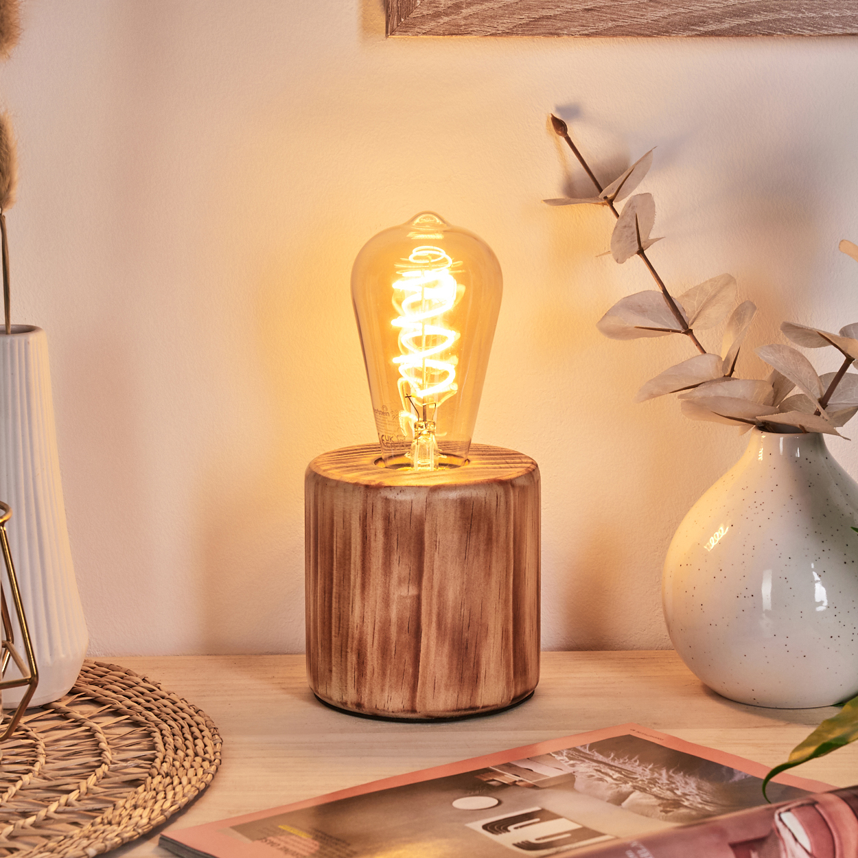 Eine beleuchtete Edison-Glühbirnenlampe auf einem Holzsockel. Vasen mit Pflanzen und eine Zeitschrift sind im Hintergrund. Die Wand ist hellbeige.