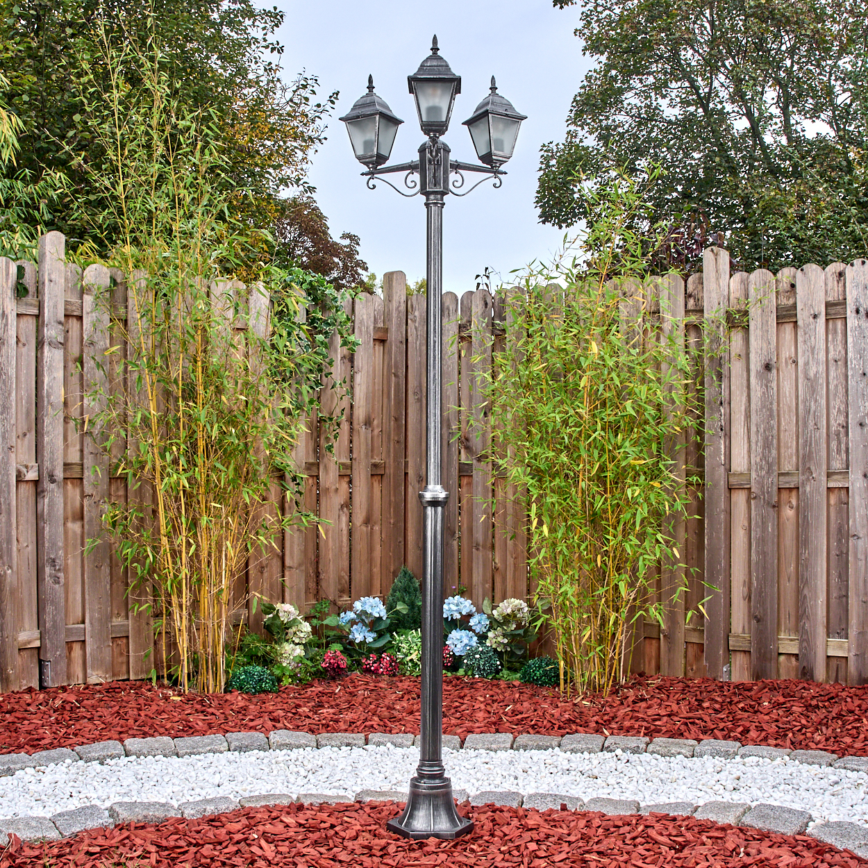 Eine dekorative Laterne steht in einem Garten mit Holzzaun. Bambus und Blumen umgeben sie, mit roter Mulch und Steinweg.