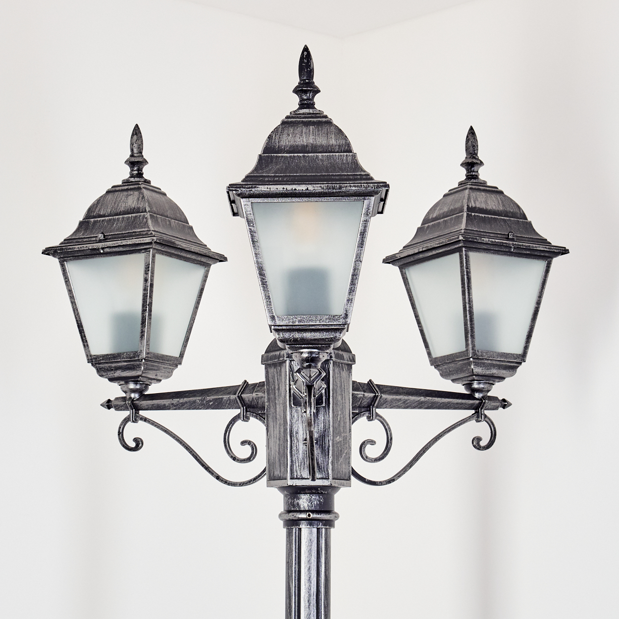 Eine dekorative Straßenlaterne mit drei Lampen aus Milchglas. Das Metall ist grau mit Zierdetails, vor weißem Hintergrund.