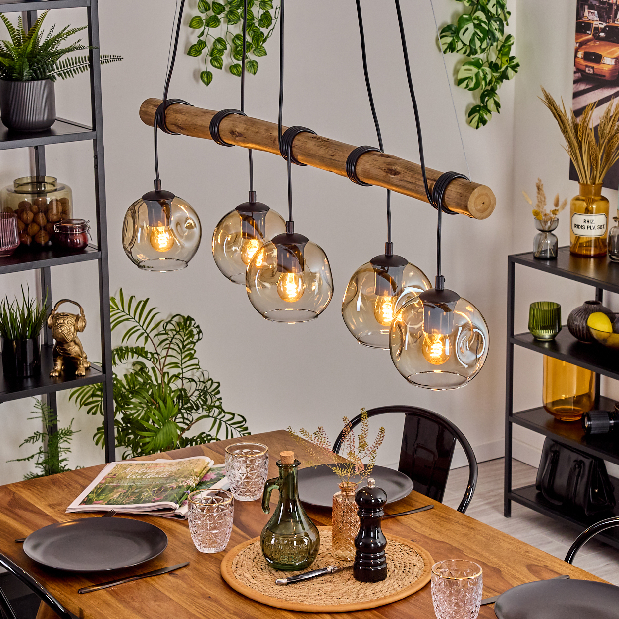 Esszimmer mit Holztisch, Hängelampen und Dekoration. Ein Holzbalken mit Glaslampen. Teller und Gläser stehen auf dem Tisch.