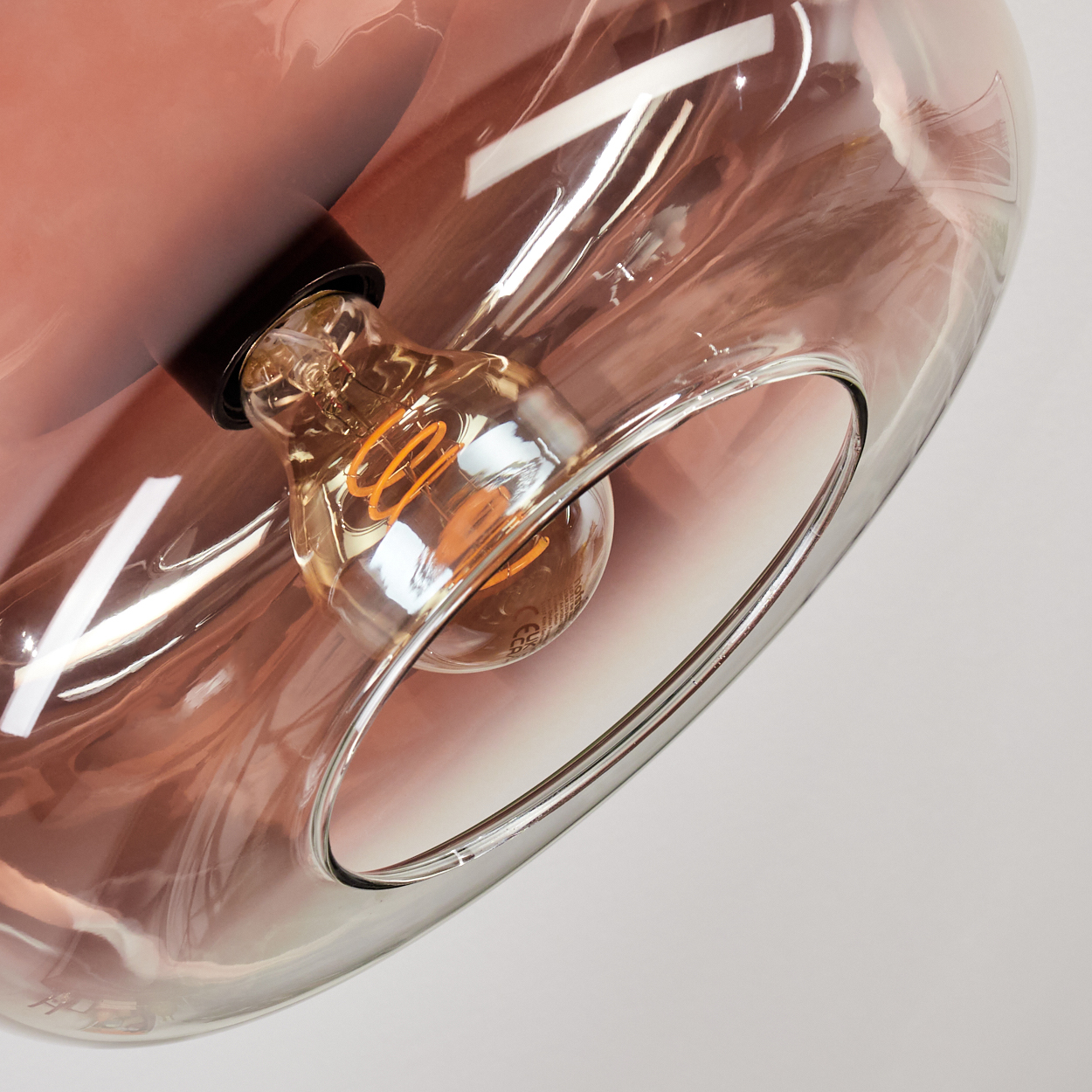 Nahaufnahme einer Glasleuchte, eine Glühbirne im Inneren mit warmem Schein. Das Glas hat einen rosa Farbton. Der Hintergrund ist weiß.