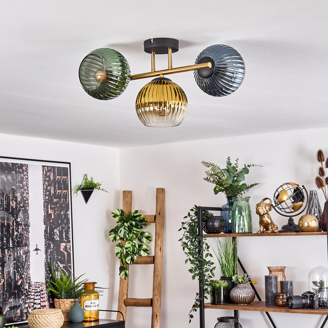 Eine moderne Deckenlampe mit drei Glasschirmen in Gold, Grün und Grau. Eine Holzleiter und Regale mit Dekor sind sichtbar.
