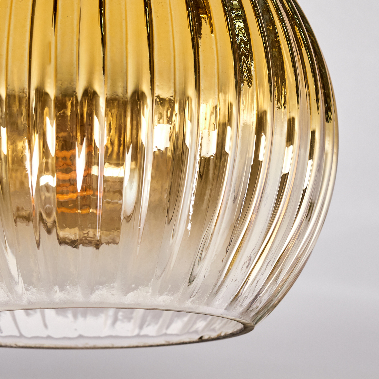Nahaufnahme eines goldenen Glasschirms. Er hat vertikale Rippen und eine Glühbirne im Inneren. Der Hintergrund ist weiß.