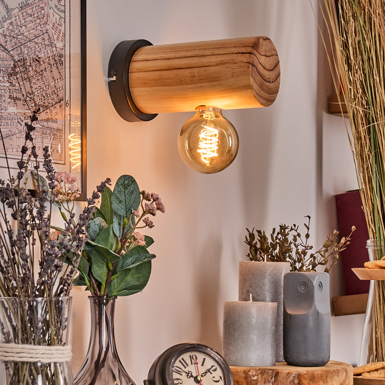 Eine Holzlampe mit leuchtender Glühbirne. Vasen mit Blumen, Kerzen und eine Uhr stehen darunter. An der Wand hängt eine Karte.