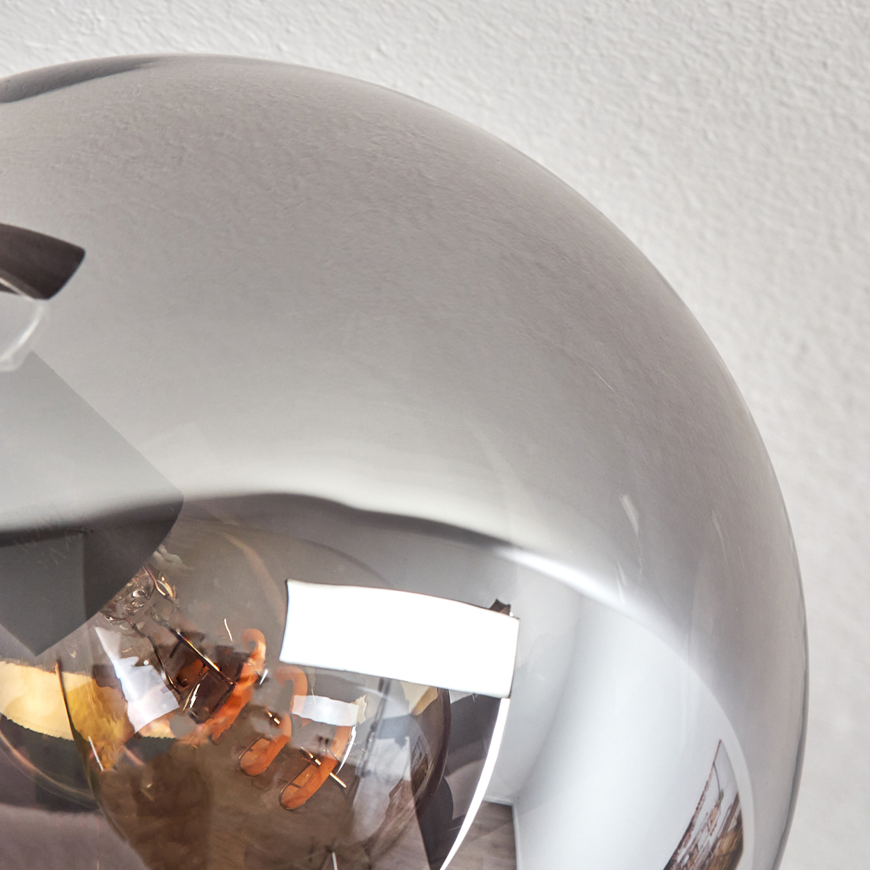 Nahaufnahme einer reflektierenden, kugelförmigen Lampe. Im Inneren der Kugel ist eine Glühbirne zu sehen. Der Hintergrund ist eine weiße Wand.