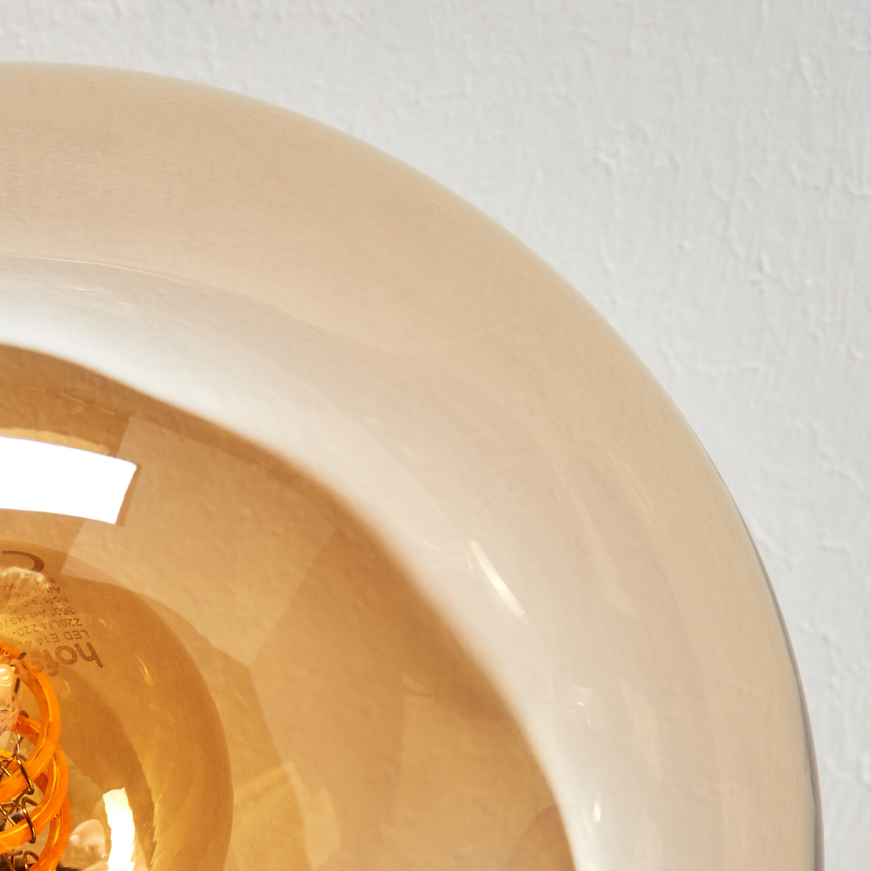 Nahaufnahme einer Leuchte. Ein beiger Glaslampenschirm im Vordergrund. Der Hintergrund ist eine weiße Wand. Die Glühbirne ist sichtbar.