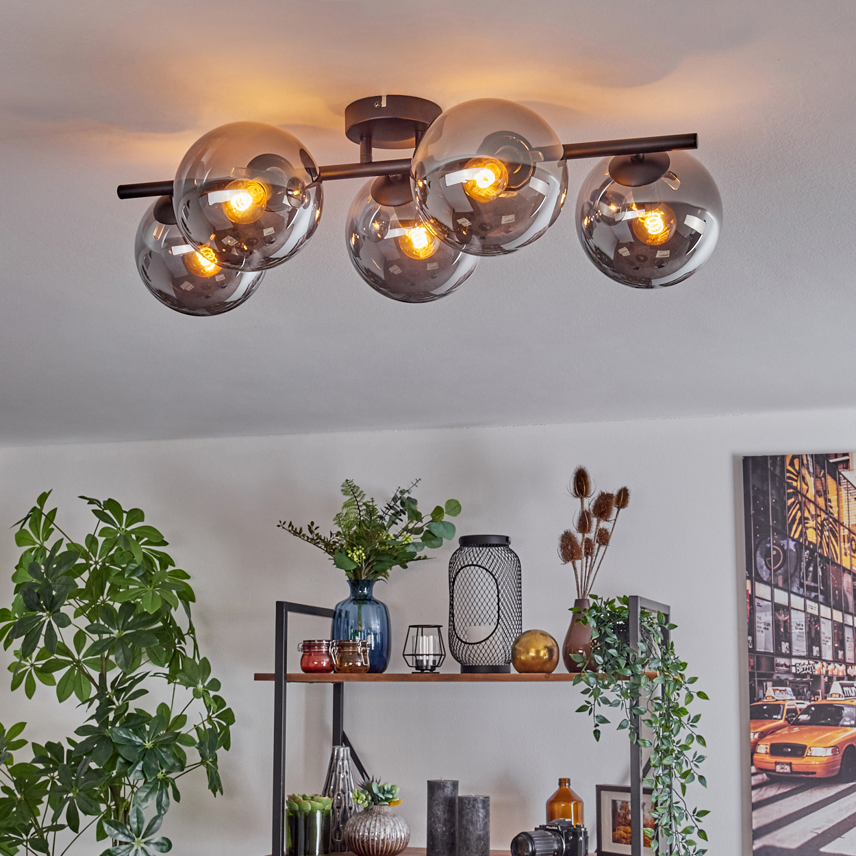Moderne Deckenleuchte mit sechs Glaskugel-Schirmen. Darunter ein Regal mit Pflanzen, Vasen, Kerzen und einem Bild vor weißer Wand.