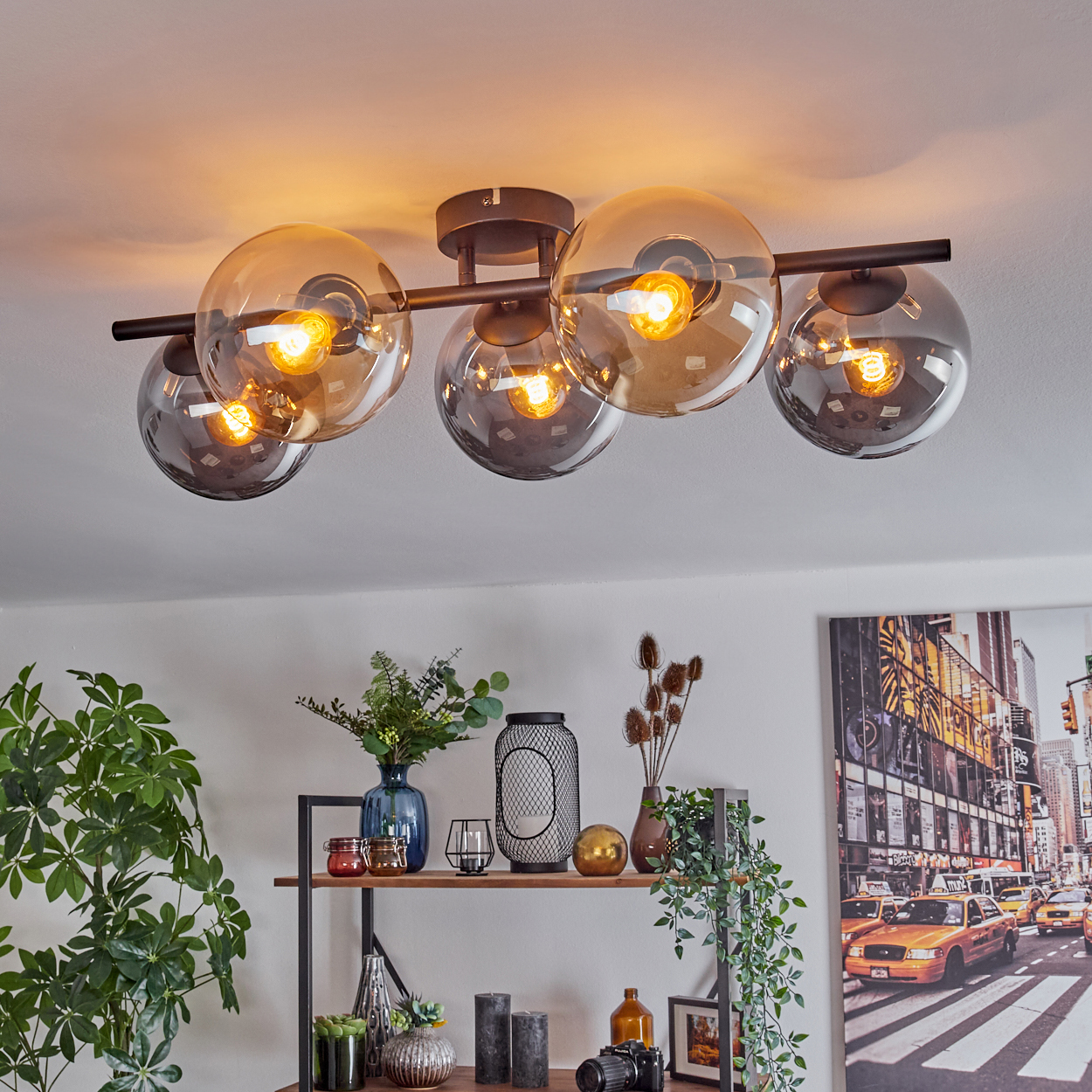 Deckenlampe mit Glaskugeln beleuchtet einen Raum. Regal darunter mit Deko, ein Stadtbilddruck rechts. Grüne Pflanze links.
