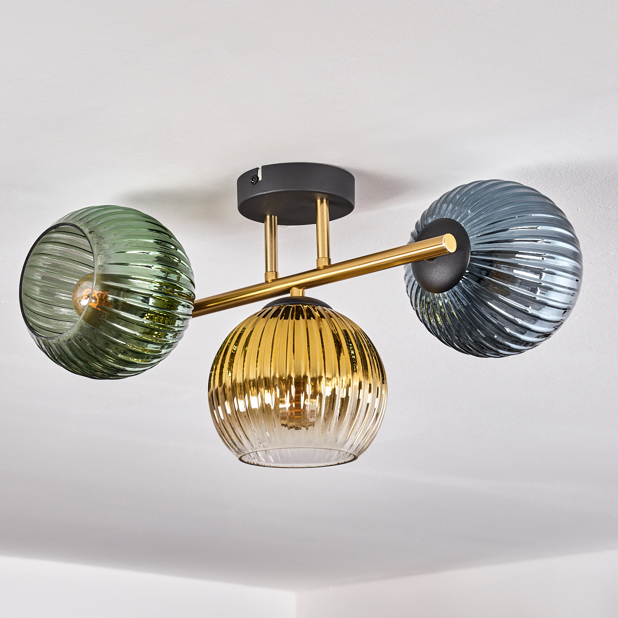 Eine moderne Deckenleuchte mit drei Glaskugeln in Grün, Gold und Grau. Die Leuchte ist an einer weißen Decke montiert.