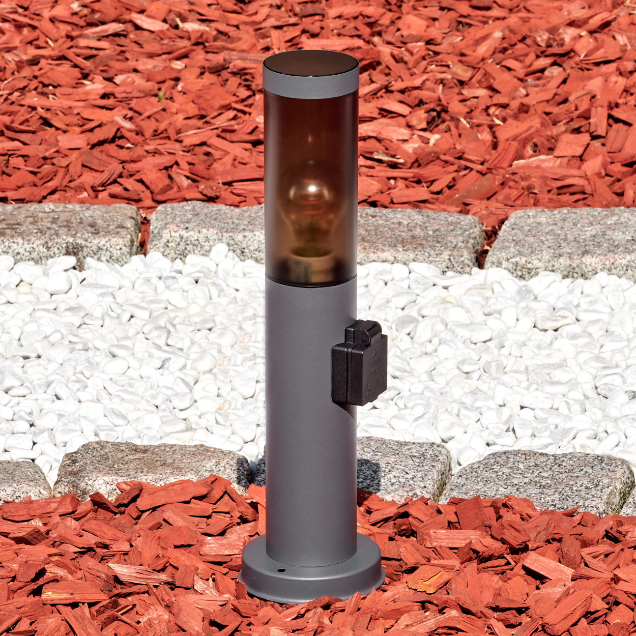 Eine graue Außenlampe mit dunklem Schirm steht auf roter Mulch und weißen Steinen. Eine kleine schwarze Box ist an der Lampe befestigt.