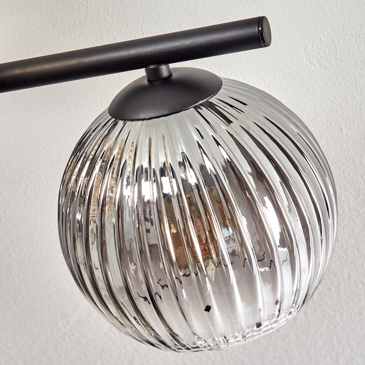Nahaufnahme einer modernen Leuchte. Sie hat einen kugelförmigen, geriffelten Glasschirm und einen schwarzen Metallarm vor einer weißen Wand.