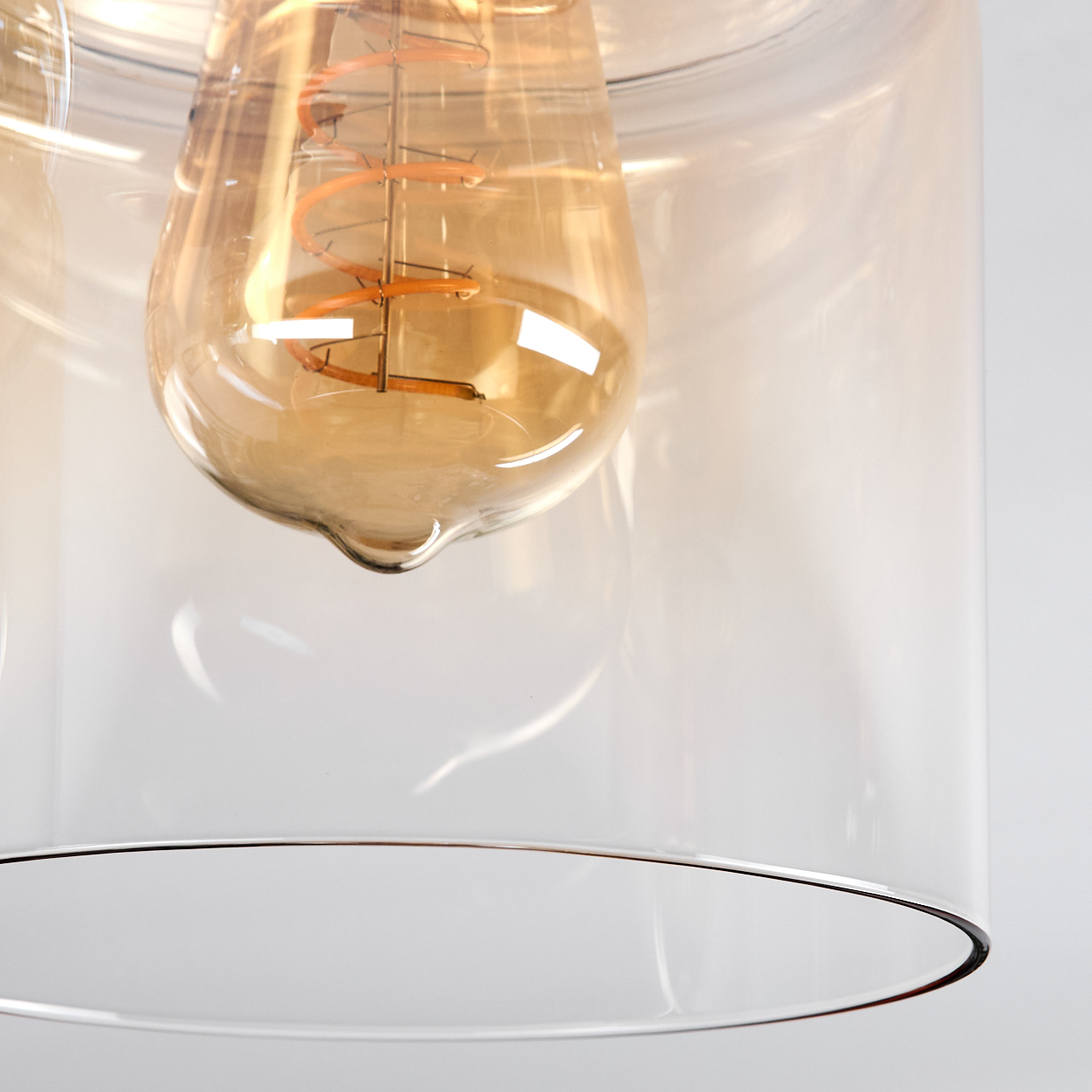 Nahaufnahme einer Leuchte mit einer tropfenförmigen, bernsteinfarbenen Glühbirne in einem klaren Glasschirm. Der Hintergrund ist weiß.