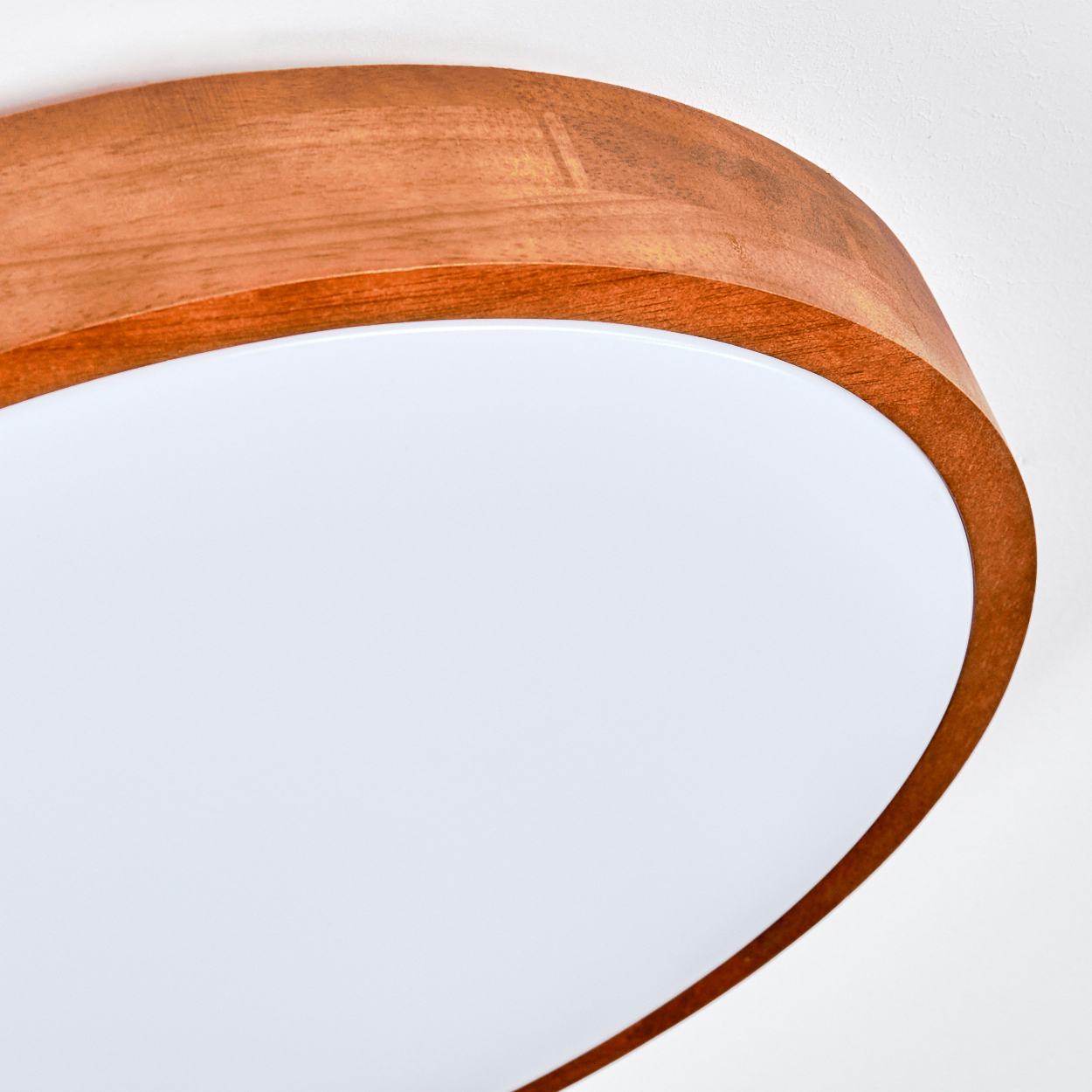Nahaufnahme einer Deckenleuchte. Sie hat einen Holzrahmen und einen weißen, abgerundeten Diffusor. Der Hintergrund ist eine schlichte weiße Wand.