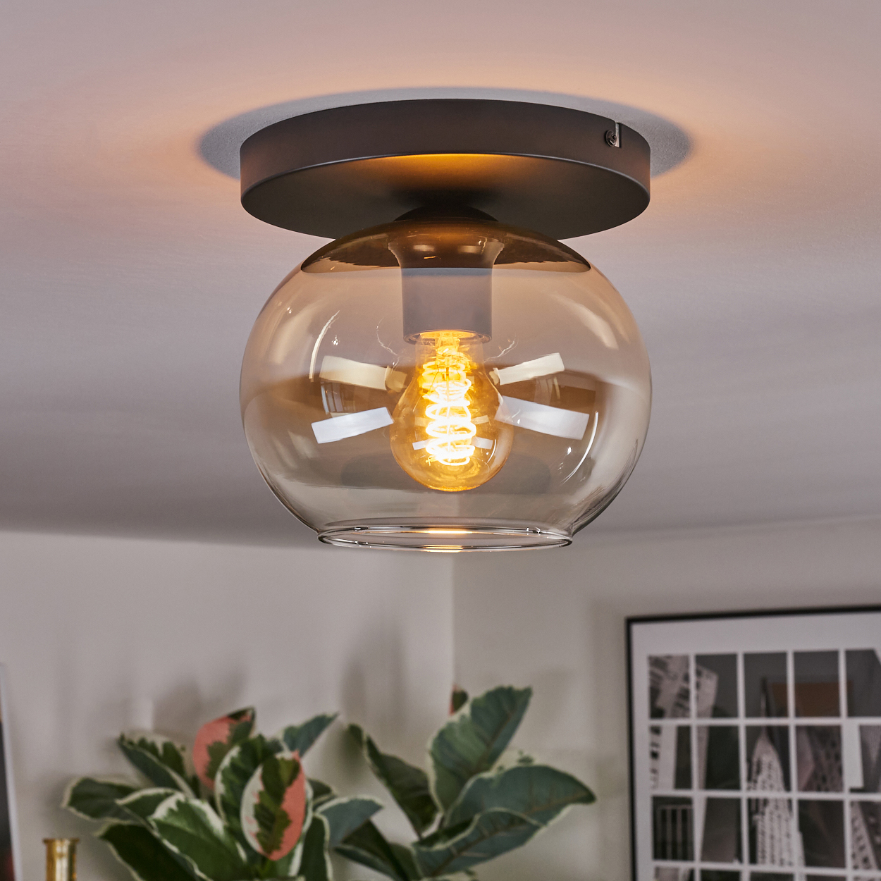 Eine moderne Deckenleuchte mit Glaskugel. Das Licht hat einen warmen Schein. Pflanzen sind im Vordergrund zu sehen. Ein gerahmtes Bild ist im Hintergrund.