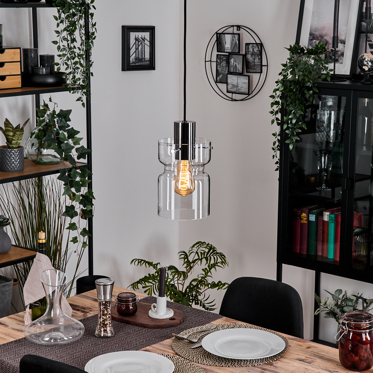 Esszimmer mit Holztisch, Glaslampe und Pflanzen. Teller und Besteck sind gedeckt. Bücherregal im Hintergrund.