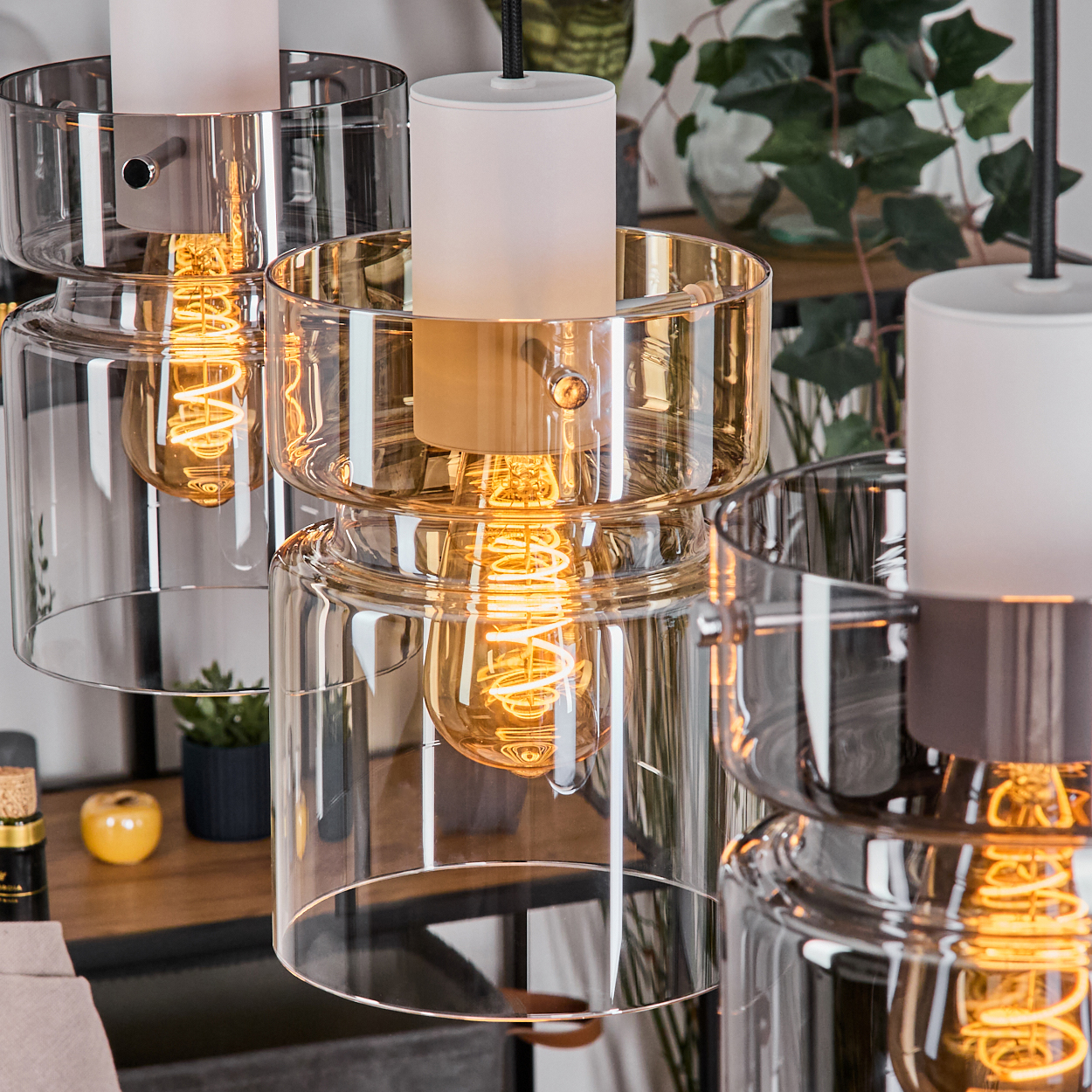 Nahaufnahme von drei modernen Glas-Pendelleuchten. Jede hat eine warme Glühbirne und einen weißen Aufsatz. Im Hintergrund ein Regal mit Deko.