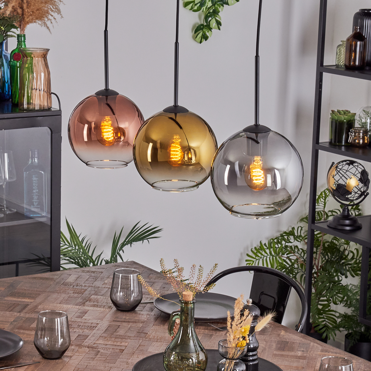 Drei Pendelleuchten mit Glasschirmen hängen über einem Holztisch, der zum Essen gedeckt ist. Pflanzen und Dekoration umgeben die Szene.