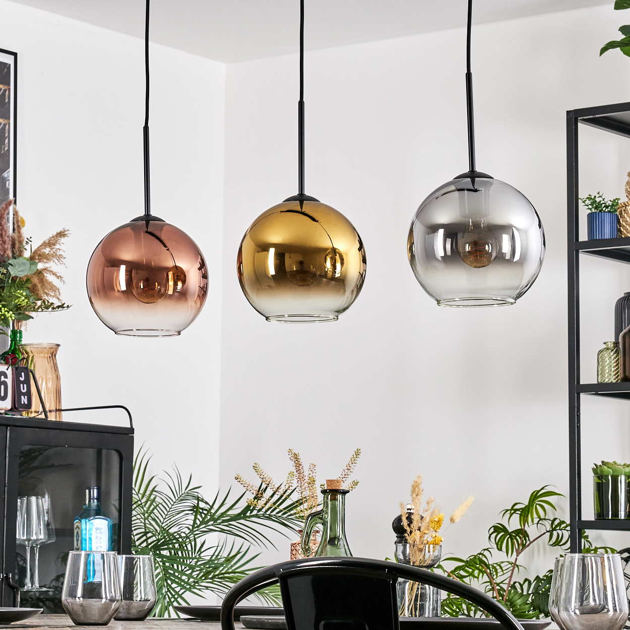 Drei Pendelleuchten mit metallischen Kugeln hängen über einem Esstisch. Die Kugeln haben Kupfer-, Gold- und Silbertöne.