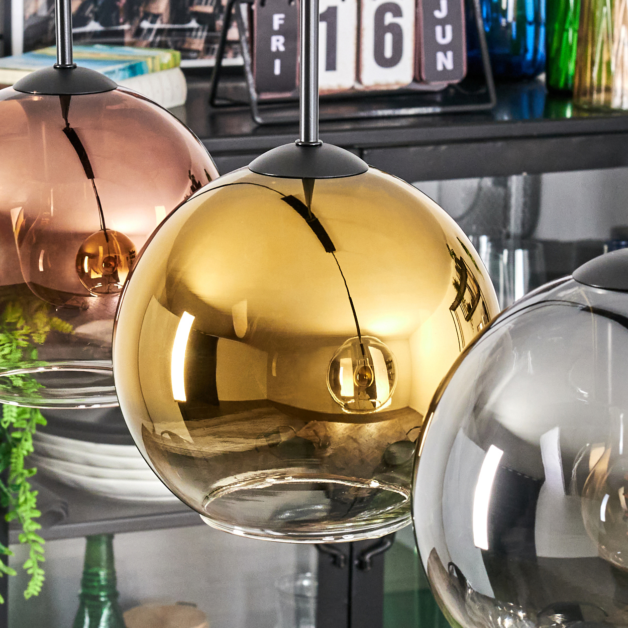 Drei kugelförmige Pendelleuchten in Kupfer, Gold und Silber. Ein Kalender und Flaschen sind im Hintergrund. Die Leuchten sind aufgehängt.