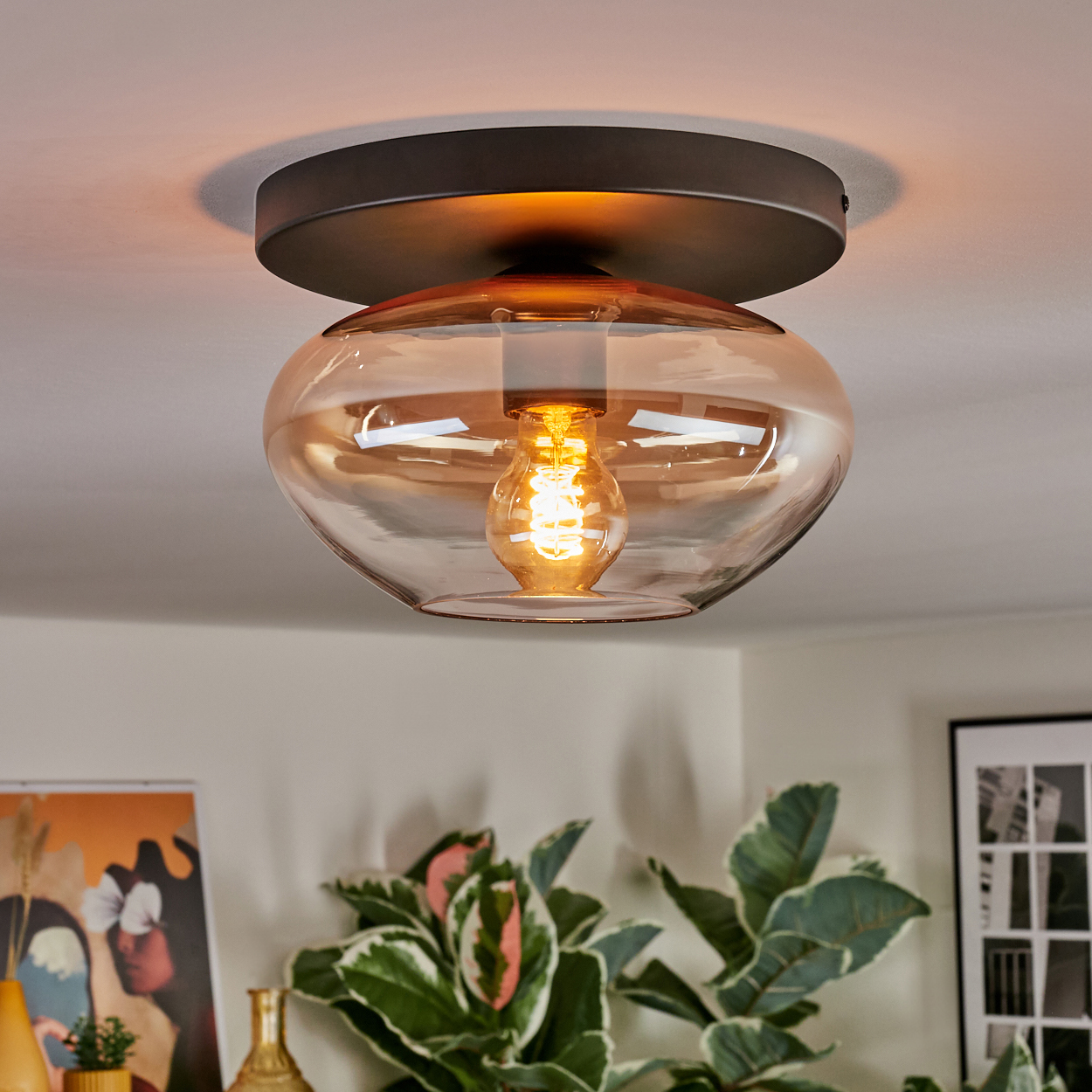 Eine moderne Deckenleuchte mit braunem Sockel und bernsteinfarbenem Glasschirm. Im Inneren hängt eine leuchtende Glühbirne. Pflanzen und Kunstwerke sind im Hintergrund.