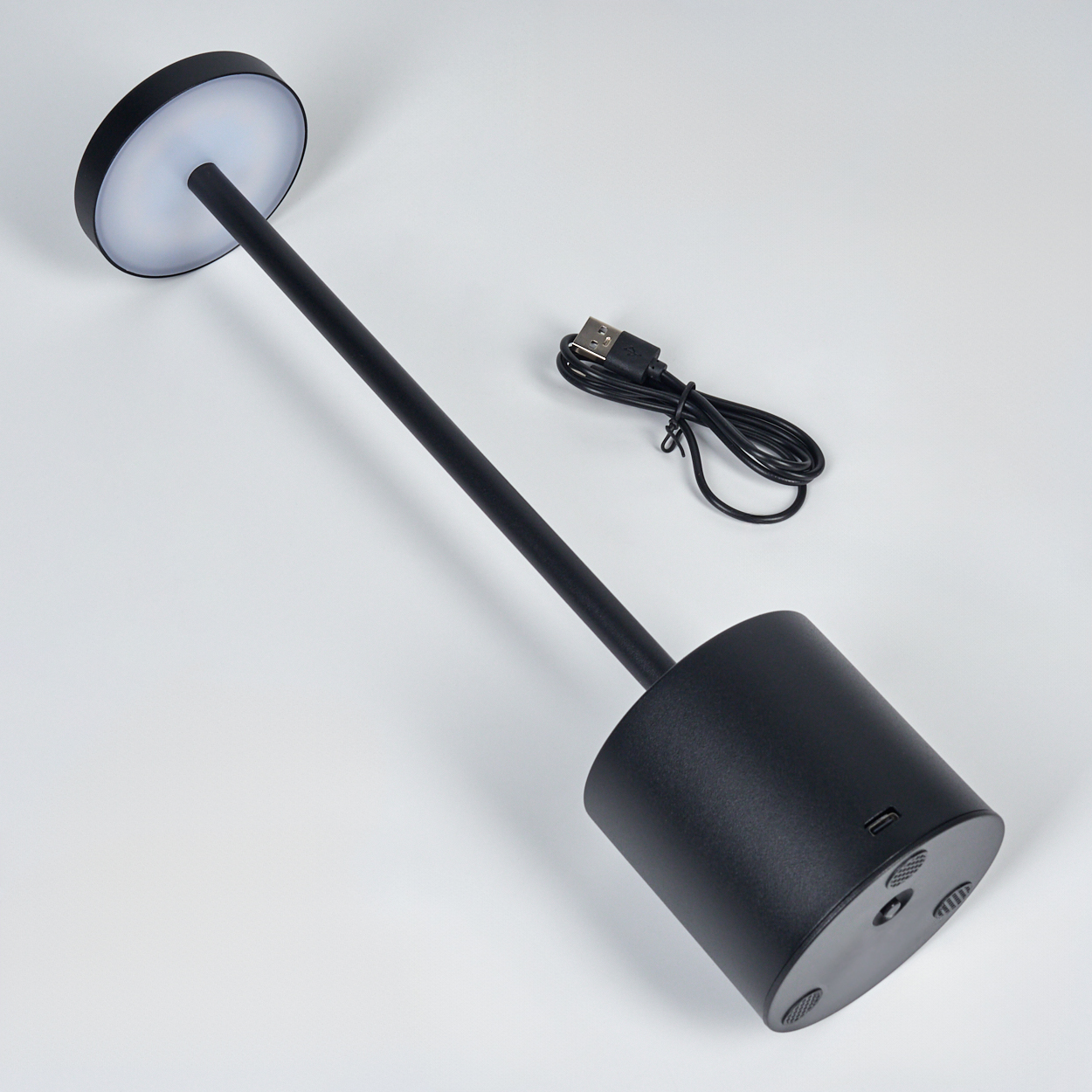 Schwarze Schreibtischlampe mit rundem Oberteil und zylindrischem Sockel sowie einem USB-Kabel, vor weißem Hintergrund.