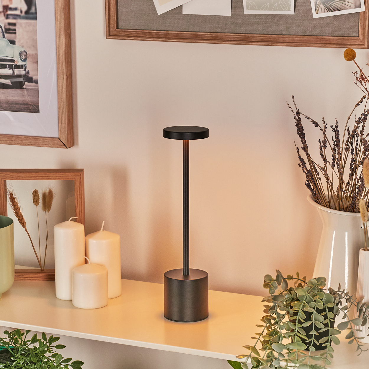 Eine moderne schwarze Lampe steht auf einem weißen Regal. Kerzen, Trockenblumen und gerahmte Kunstwerke sind in der Nähe. Grüne Pflanzen im Vordergrund.