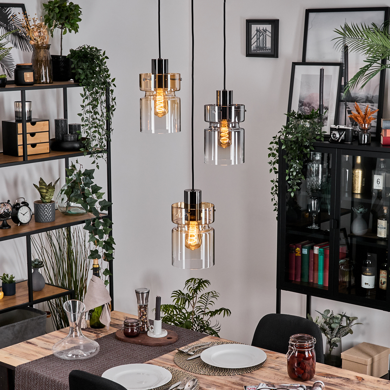 Esszimmer mit Holztisch, drei Hängelampen, schwarzen Regalen mit Dekor und einem schwarzen Schrank mit Büchern und Gläsern.