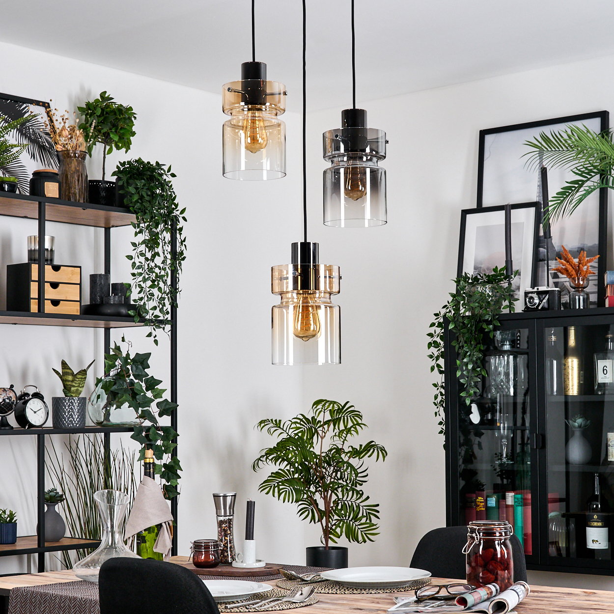 Esszimmer mit Hängelampen, Pflanzen und einem schwarzen Schrank. Tisch mit Tellern und Gläsern. Gemütlicher, möblierter Raum.