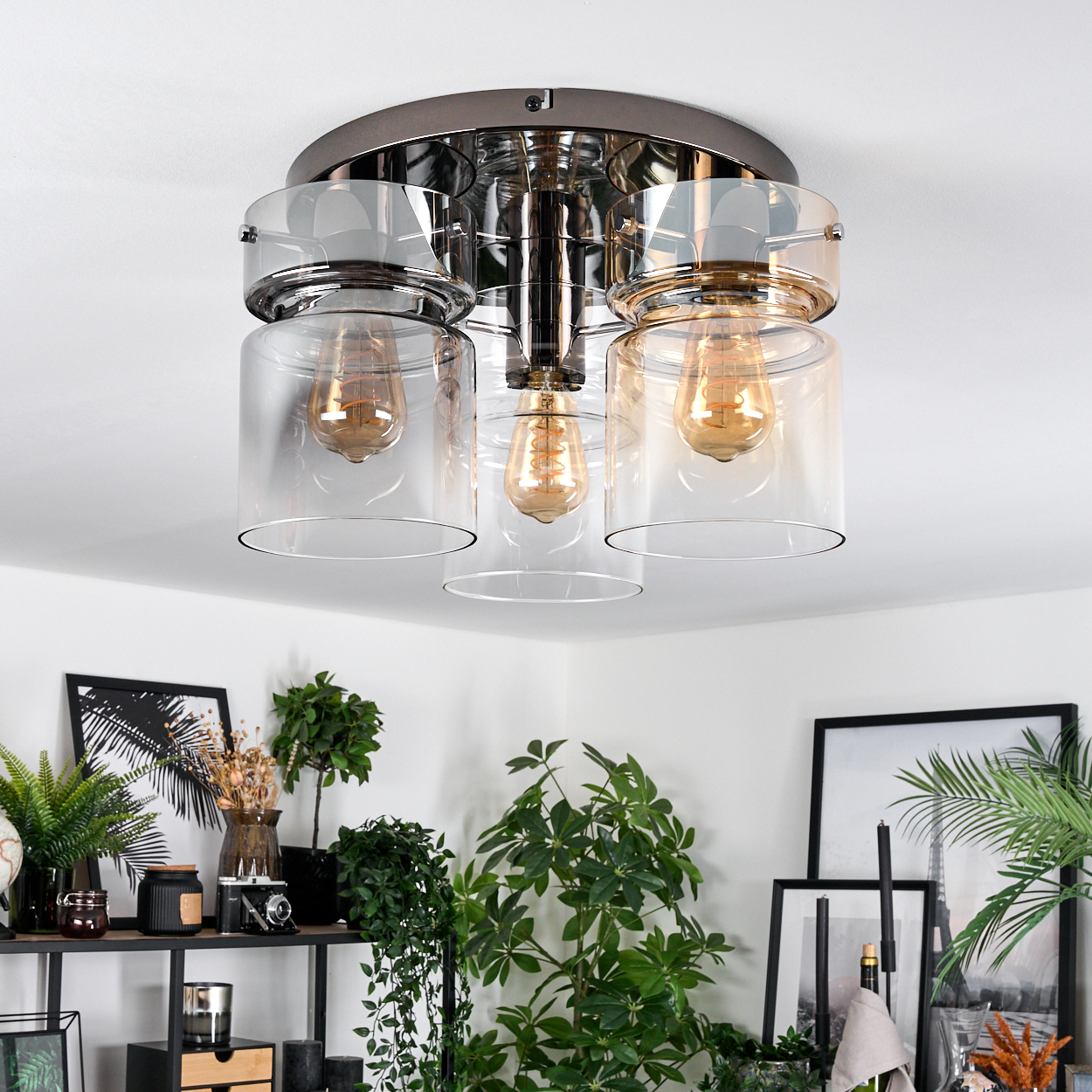 Eine moderne Deckenlampe mit drei Lichtern. Der Raum hat Pflanzen, Bilder und ein Regal mit Objekten.