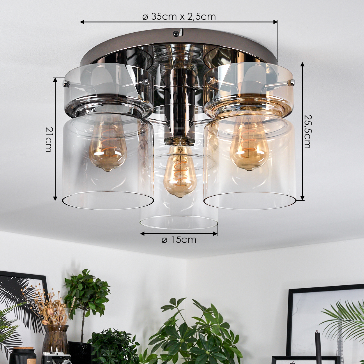 Eine moderne Deckenlampe mit drei Glasschirmen, die jeweils eine Glühbirne enthalten. Abmessungen sind angegeben. Pflanzen und gerahmte Kunstwerke sind im Hintergrund.