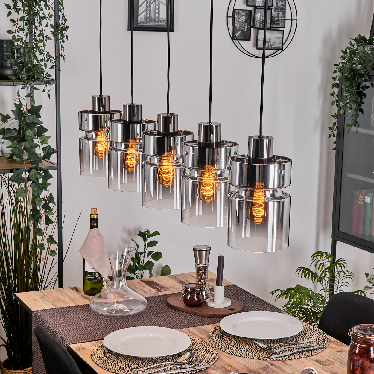Esszimmer mit Holztisch. Fünf Pendelleuchten hängen darüber. Teller, Wein und Pflanzen sind sichtbar. Wanddekor beinhaltet Fotos.