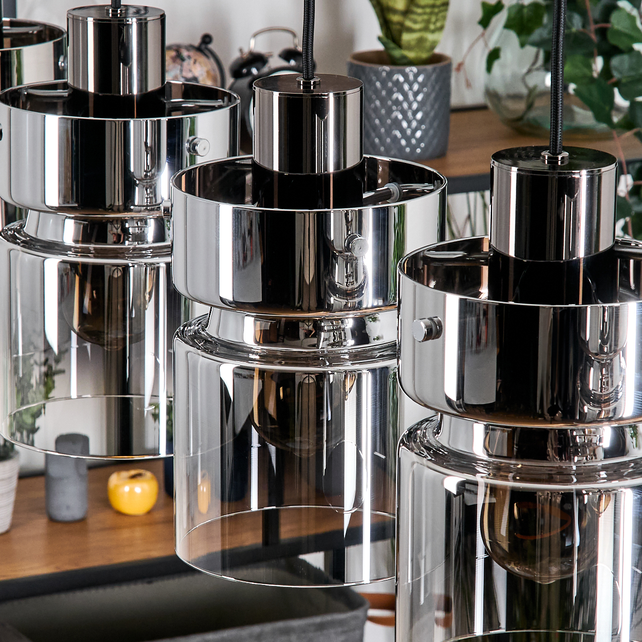 Nahaufnahme von drei modernen Pendelleuchten mit Chrom-Finish und Glasschirmen. Im Hintergrund Pflanzen und ein Holzregal.